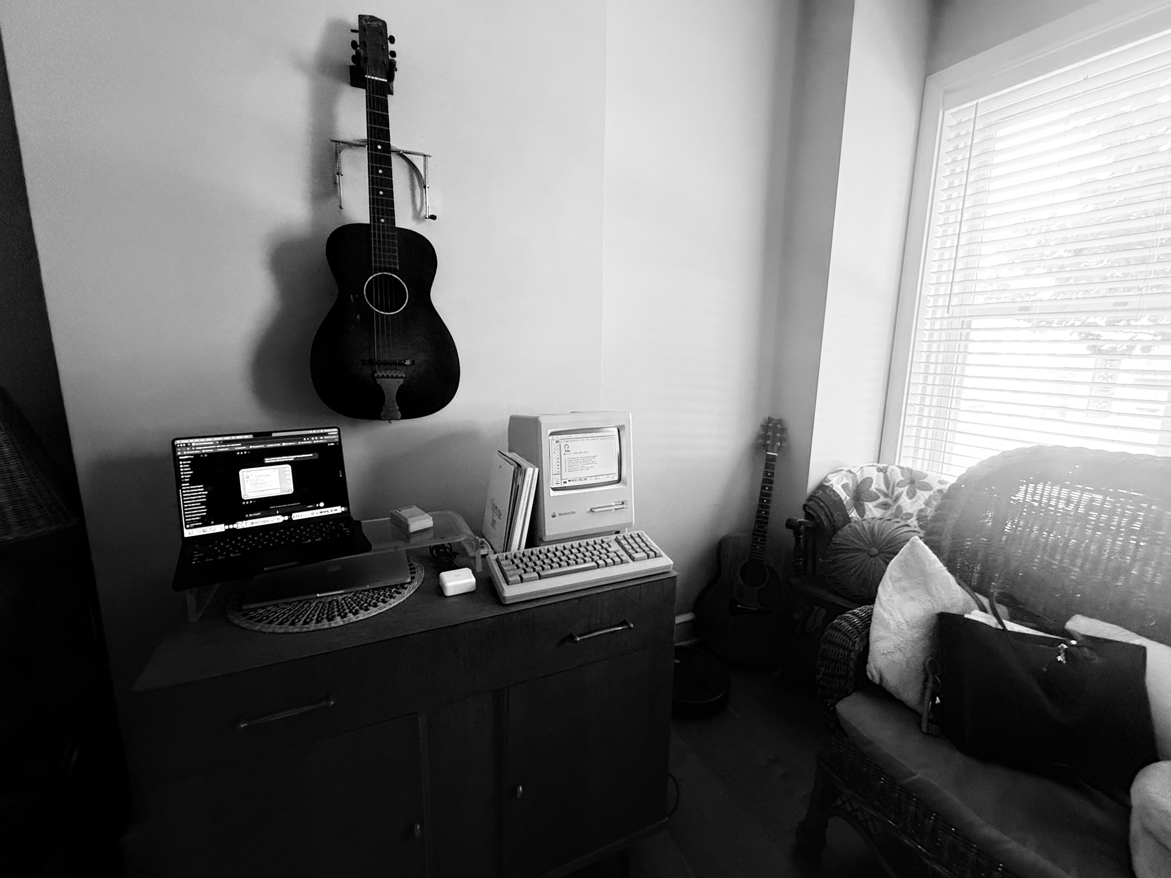 A real Macintosh Plus sitting on a desk next to a modern laptop, under a vintage guitar